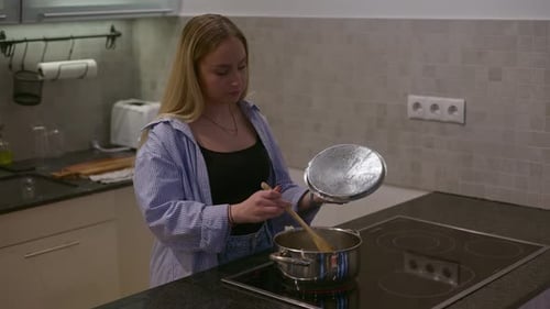 A young woman is standing at the stove in a modern kitchen, stirring food in a stainless steel pot