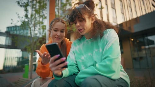 Two Young And Stylish Female Students Talking On City Street And Using Smartphone To Scroll Social