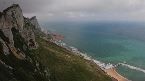 A dramatic aerial view of a rugged coastal cliff descending into a turquoise sea. The shoreline