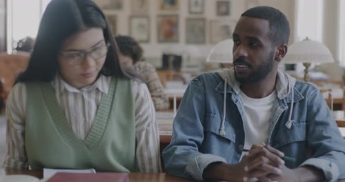 Portrait of Young Asian Woman Sitting Down at Table in College Library and Talking to African