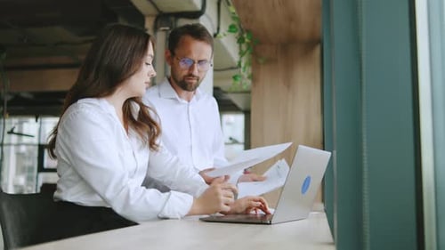 Business Team Discusses Work Tasks While Sitting at a Table Near a Large Window in a Modern Office