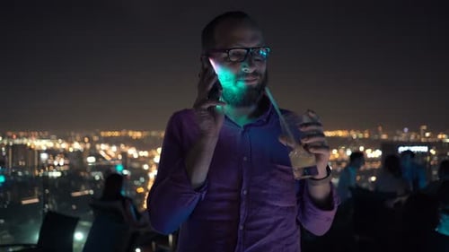 Handsome young man chatting on cellphone and enjoying cocktail at city bar terrace night