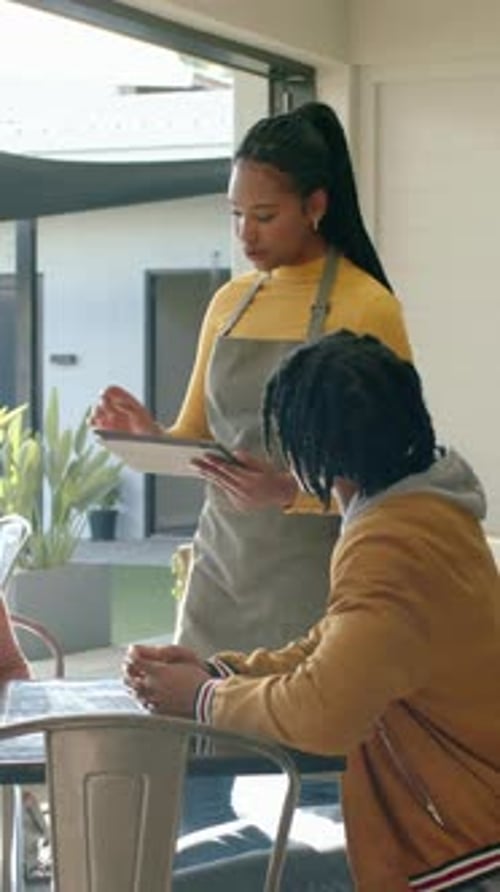 Vertical video: Approaching server in yellow top, gray apron showing tablet, taking order at table