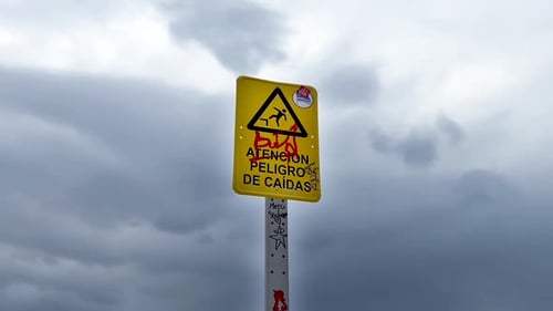 grafitti on spanish public warning sign next to the sea, cloudy sky.
