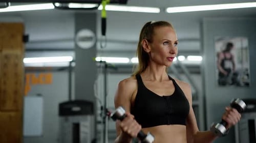 A Young Girl Athlete with Confident Look in the Gym Does Exercises with Dumbbells Breathes Raises