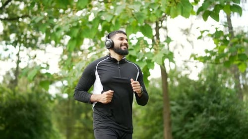 Adult bearded male runner in wireless headphones jogging outside in urban city park. Handsome man