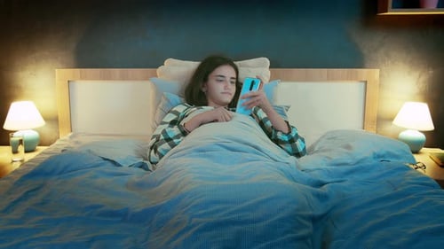 Girl Lying Under the Covers at Night in Her Bed and Browsing Mobile Apps Front View