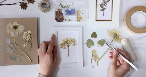 Woman putting dry flowers into picture frame at white marble table, top view
