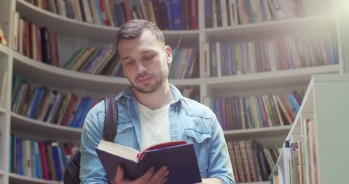 Young Handsome Caucasian Male Student in with Backpack Standing in Library Reading Textbook and