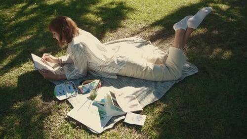 An Adult Inspired Woman Lying on the Blanket in the Park and Drawing in Her Sketchbook