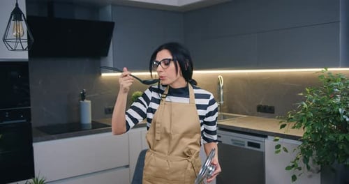 Woman in Apron Tastes Food in Modern Kitchen