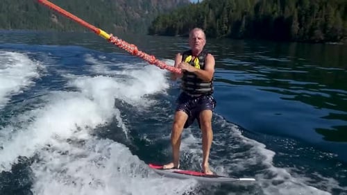 Older man wake boarding on beautiful lake striking a pose and showing off biceps.