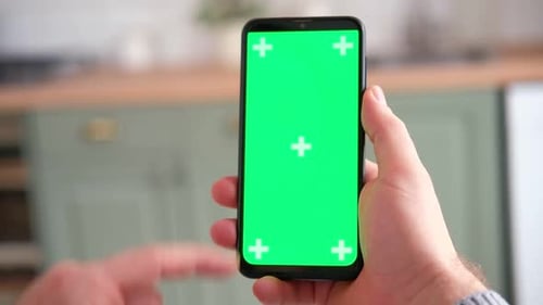 Young Man Sitting at Home Holding Smartphone Green Mockup Screen in Hand