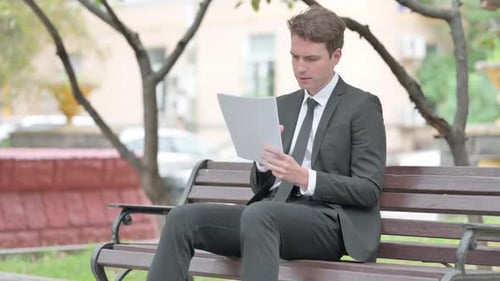 Man in Suit Reading Documents in Park Looking Distraught