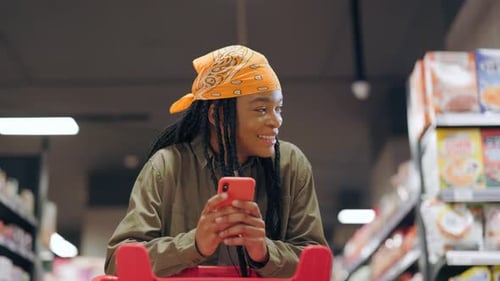 Black Attractive Cheerful Woman with Dreadlocks Inside Supermarket Chatting Messages in Smartphone