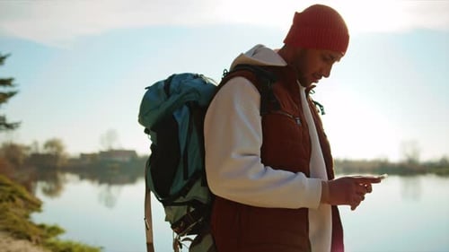 Side View of Man with Tourist Backpack on Background of Lake Using Smartphone