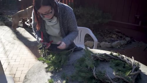 Young woman wearing sweater clips pine branches for a wreath while a yellow labrador retriever stand