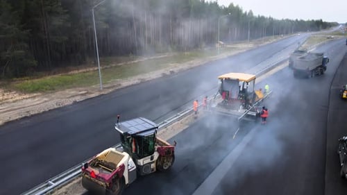 Road roller compacts the hot asphalt after paver in puffs of steam, drone view