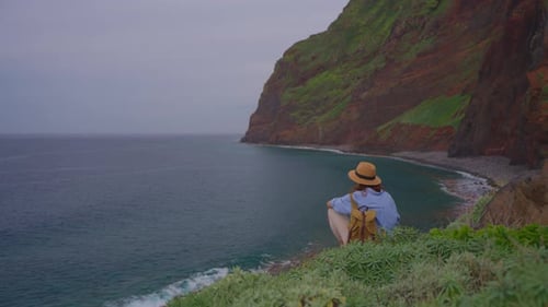Girl Traveler Sitting on a Rock in a Hat and with a Backpack Enjoying the View of the Ocean