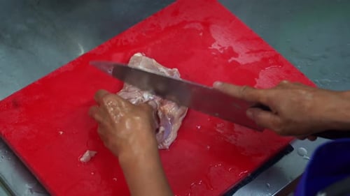Ansicht von oben, die Hände eines Profikochs beim Schneiden und Portionieren von rohem Hähnchenschenkelfilet auf einer roten Platte fotografiert