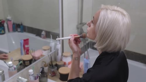 Woman Applies Makeup With Brush in Bathroom Mirror