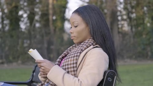 Beautiful black young woman in park sitting on bench reading book trees cloudy day central London. S