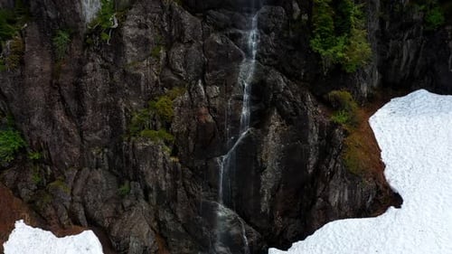 Malerische Luftaufnahme eines kleinen Bachwasserfalls, der die felsige Klippe hinunter in den Schnee fließt.