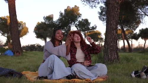 Female friends enjoying picnic moment, sitting on blanket in sunlit urban park, pointing skyward and