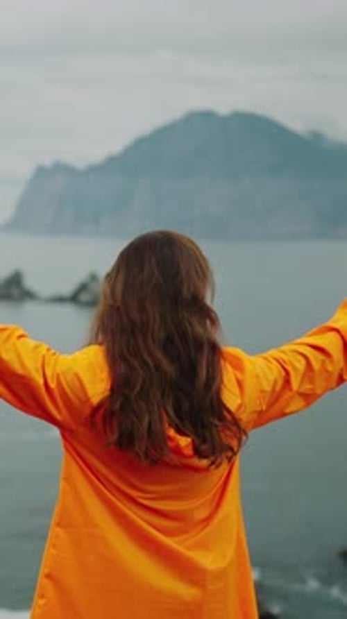 A Joyful Embrace of Nature's Majesty A Woman in an Orange Jacket Standing on the Coast with Arms