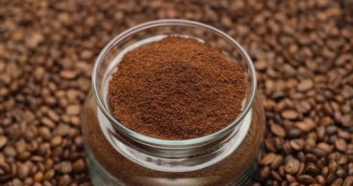 Glasschale voller gemahlener Kaffee auf einem rotierenden Hintergrund aus frisch gerösteten Kaffeebohnen