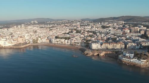 Drone flies towards coastal town lit by warm morning sun as gentle sea waves quietly roll onto the s