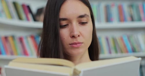 Smiling Young Girl Reading Book in Library of University and Looking at the Camera