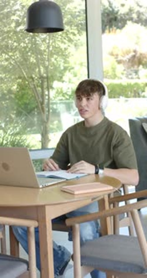 Vertical video: Teen boy in olive tee with headset joining laptop class and studying notes by wind