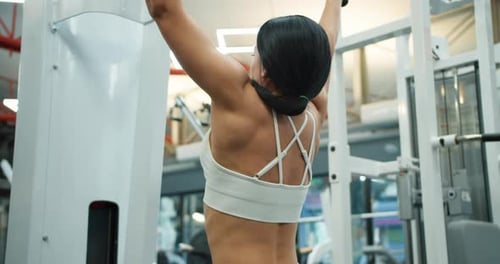 Back View of Athletic Girl in White Top Doing Back Muscle Exercises on Block Trainer in Modern Gym