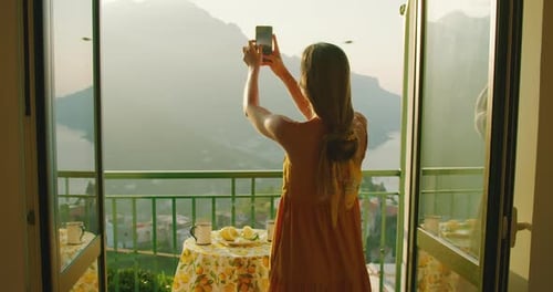A woman captures the Amalfi Coast sunrise on her smartphone, framed by a balcony setting with lemons
