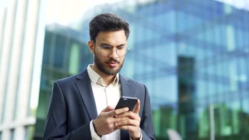 Confident businessman in suit stands near modern glass business office building holding smartphone