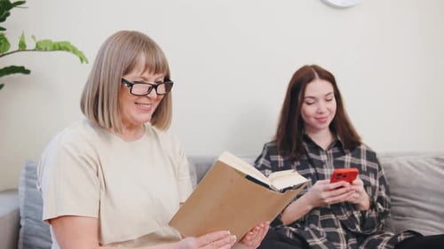 Senior Woman Reading Book with Young Adult on Phone