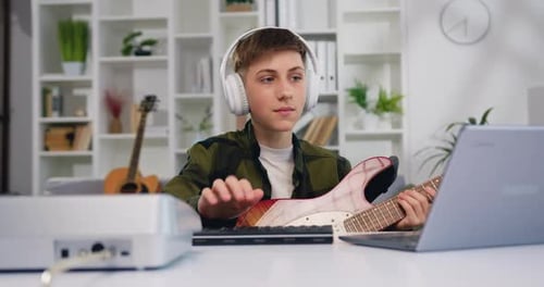Atractivo chico de 13-14 años con auriculares tocando la guitarra mirando el portátil en casa. Retrato
