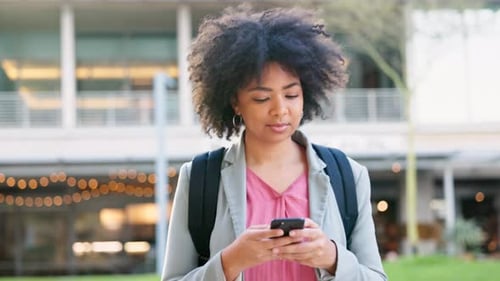 Happy female student typing on a phone and walking outside on university campus