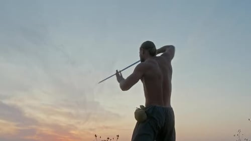 Muscular Shirtless Man with Sword Practicing Slashing Movements in a Low Angle View Against the