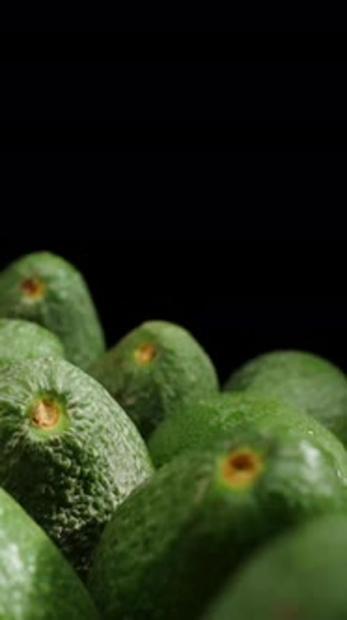 Vertical video. Camera moves backward past neatly arranged avocados on a table, some of them falling