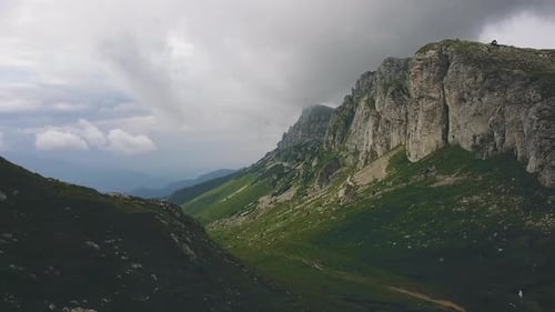 Drone backward shot revealing a mountain ridge with low clouds and an alpine valley with green grass