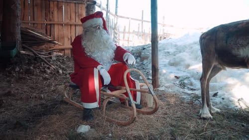 Le Père Noël à l'extérieur, près d'une maison en bois, assis sur son traîneau avec l'un de ses cerfs et discutant.