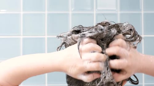 Woman washes her hair with shampoo on blue background, back view.