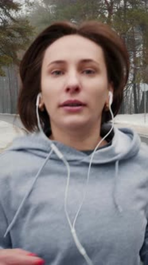 Portrait of female in earphones and sportswear jogging in park. Vertical video