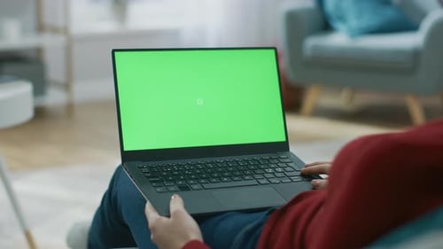 Young Woman at Home Works on a Laptop Computer with Green Mock-up Screen. She's Sitting On a Couch