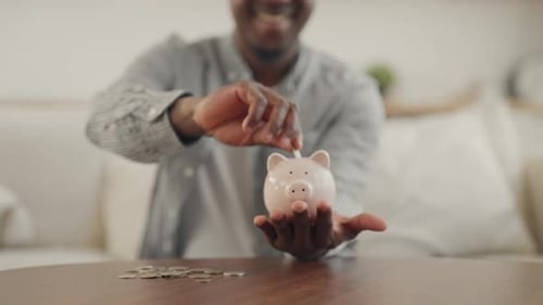 Young Adult Black Man Putting Coins in Piggy Bank Closeup of Hands at Home Saving Money and Future