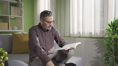 Man Sitting Indoors, Reading Book on Couch