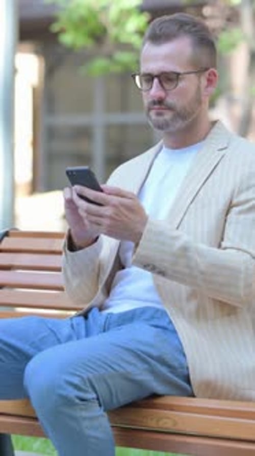 Bearded Man in Blazer Sits on Bench with Phone