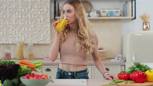 Woman Drinks Juice Amidst Vegetables in Sunny Kitchen